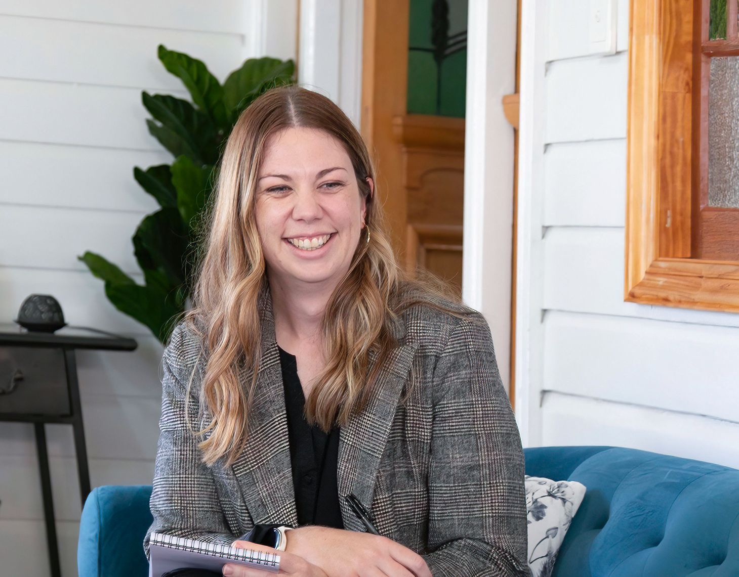 Smiling woman seated on a teal couch in a bright porch, holding a notebook