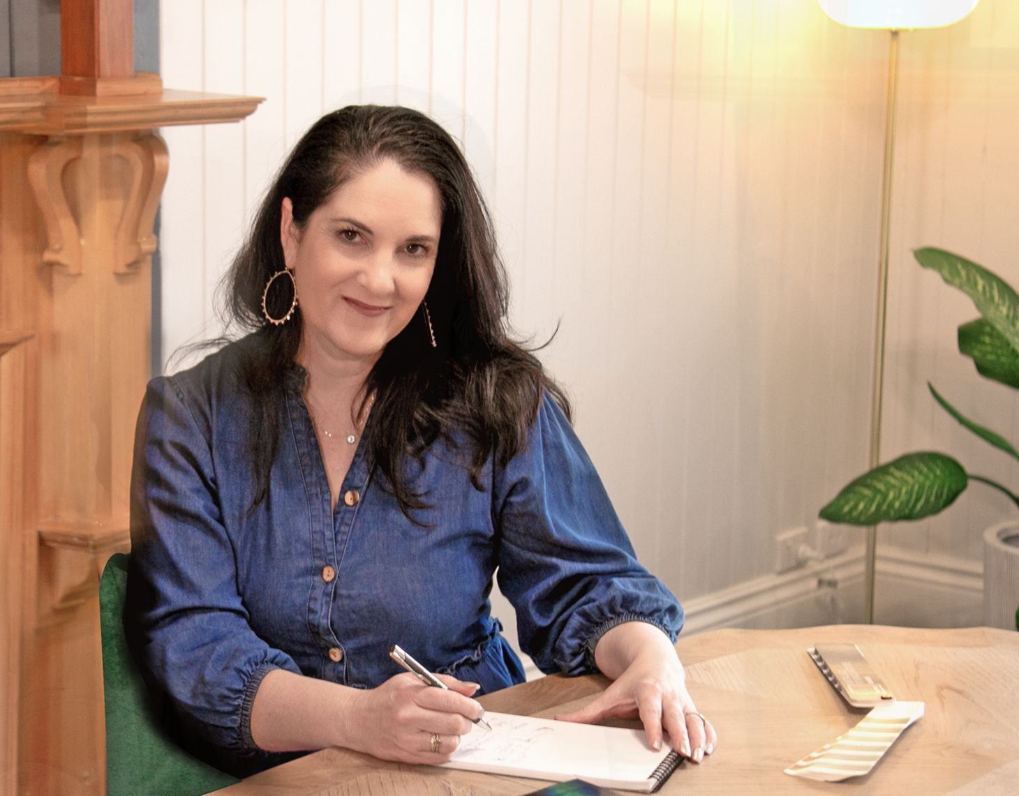 Woman writing in a notebook at a wooden table in a bright room, wearing a blue blouse.