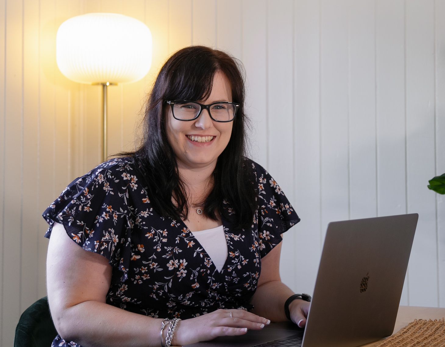 Smiling woman with glasses using a laptop beside a glowing lamp
