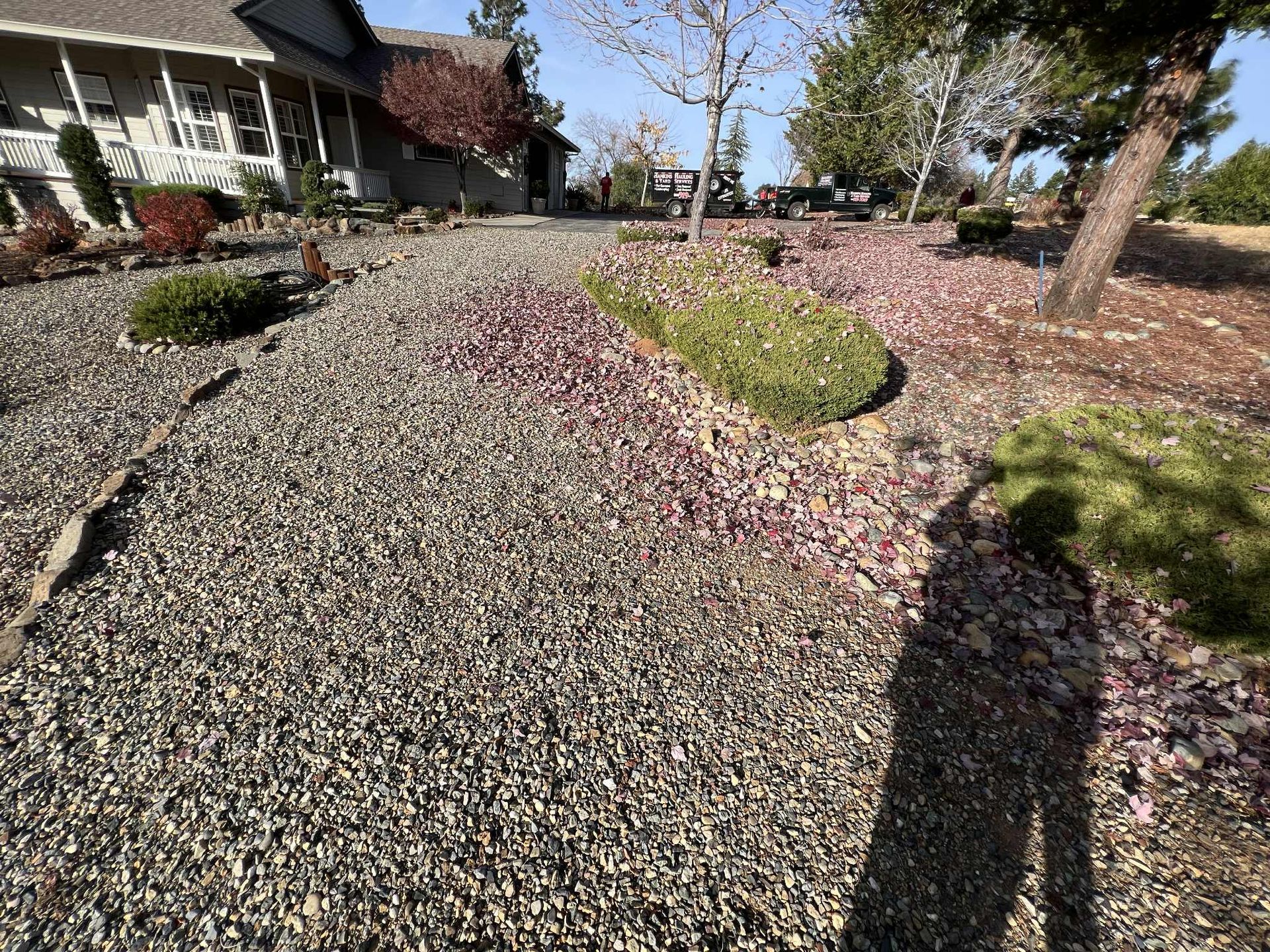 Gravel pathway leading to a house with landscaping, person's shadow in foreground, trees and bushes.