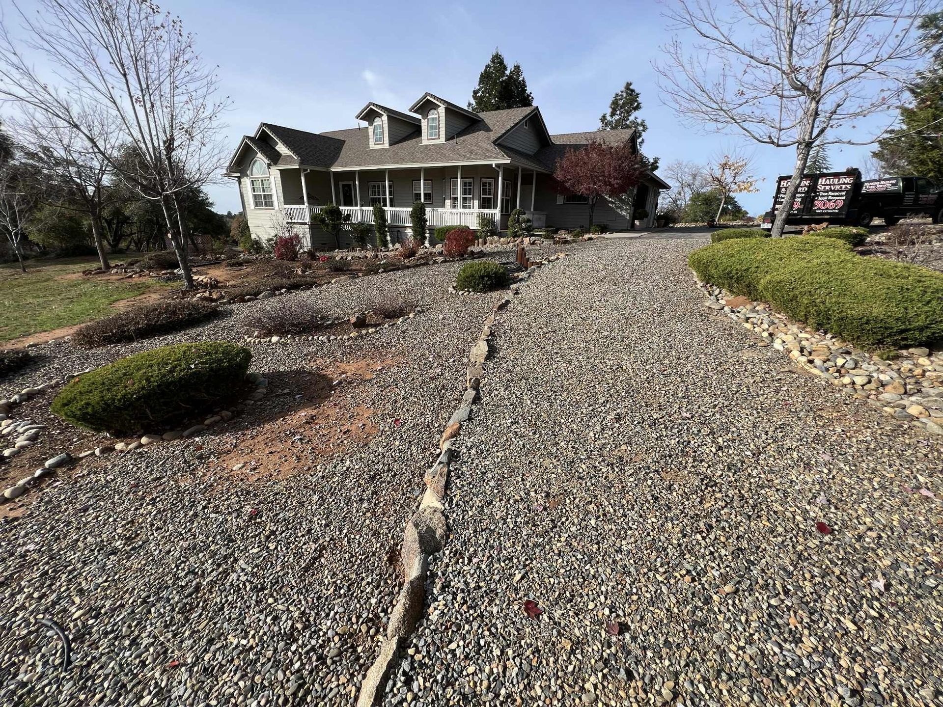 Gravel driveway leads to a two-story house with a porch, surrounded by landscaping and trees under a blue sky.