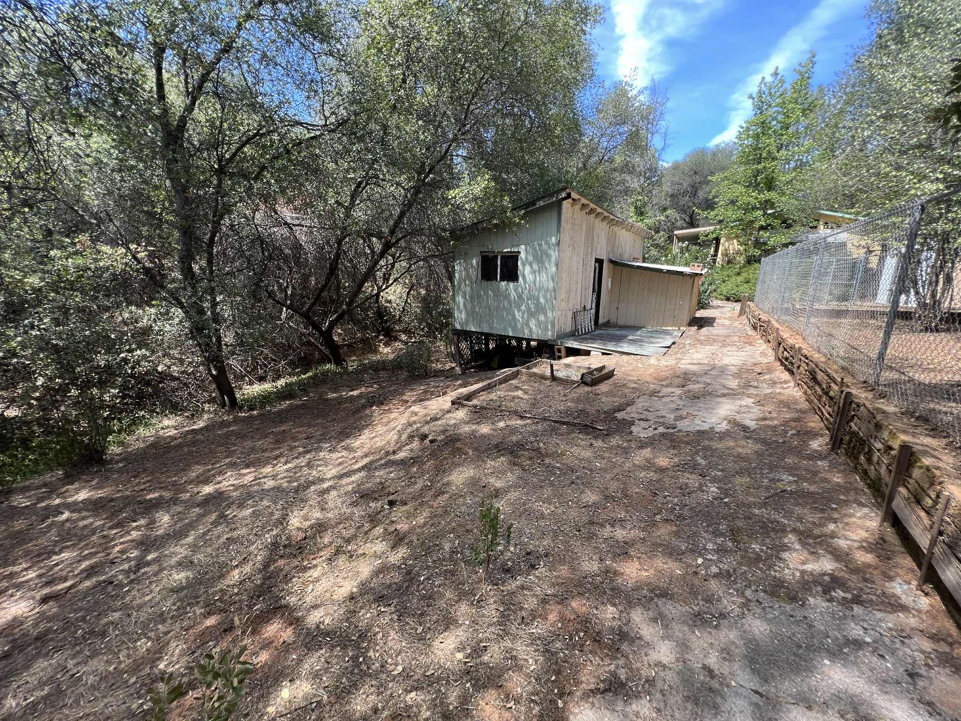 Small weathered shed in a wooded area with a dirt path leading toward it, chain-link fence on the right.