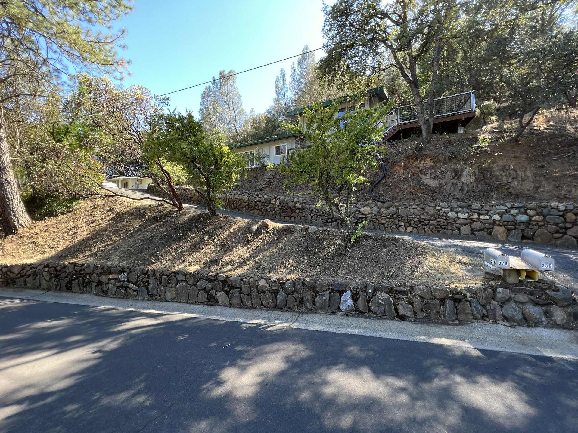 A house on a hillside with stone retaining walls, trees, and a road in front.