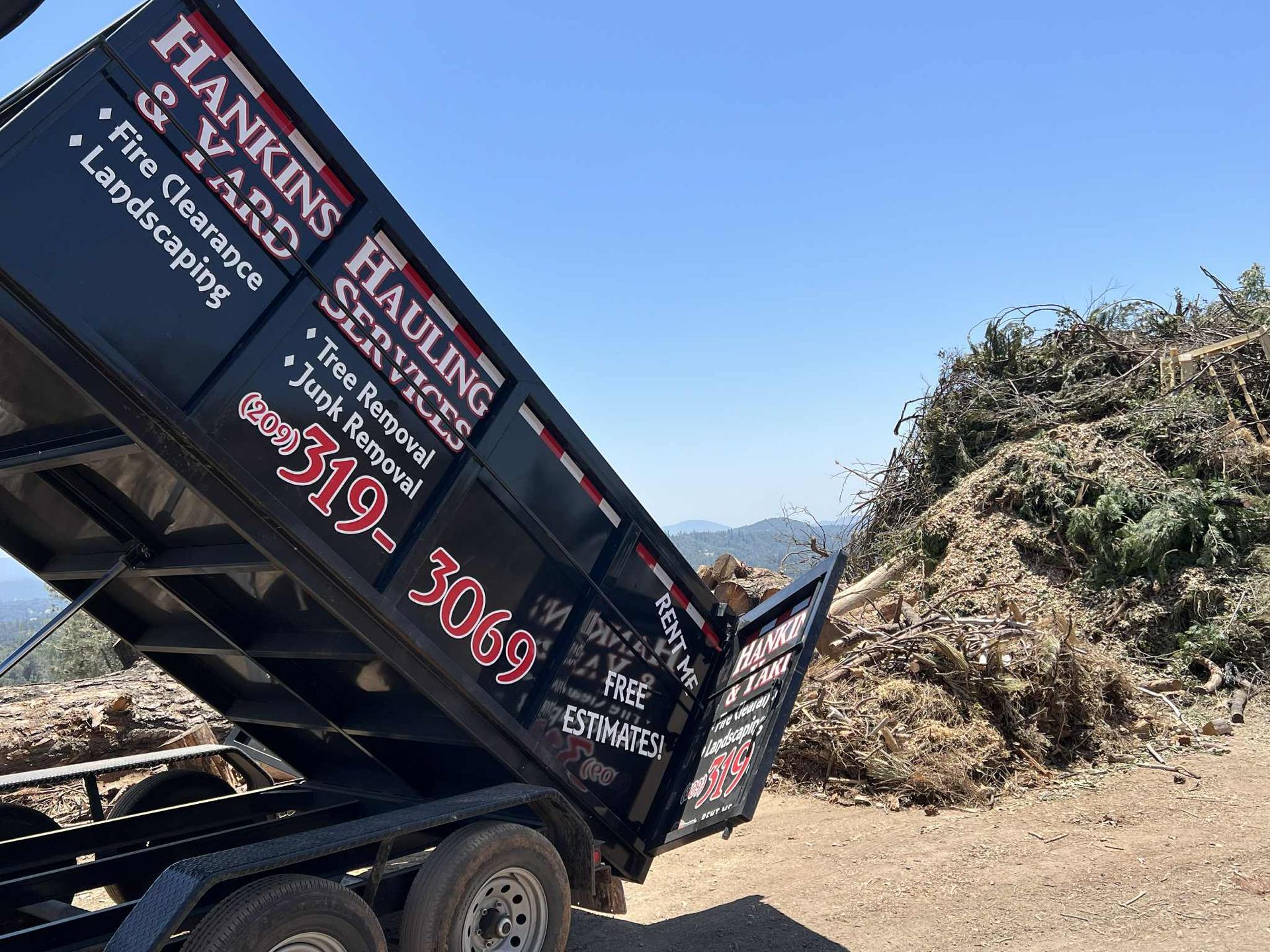 A black dump trailer unloading yard waste; blue sky in the background.