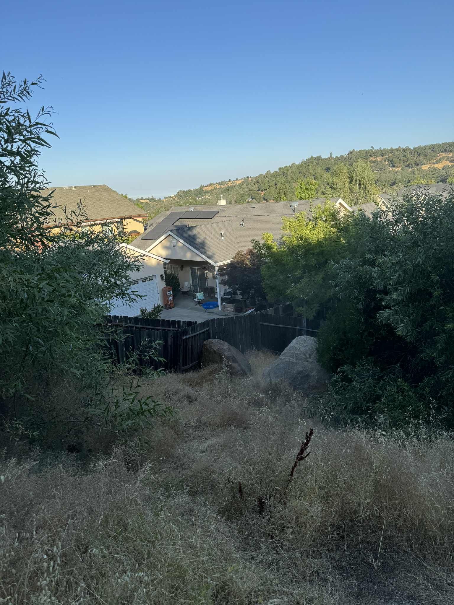 View of a house from a hill covered with dry grass and bushes, with a mountain in the background under a blue sky.