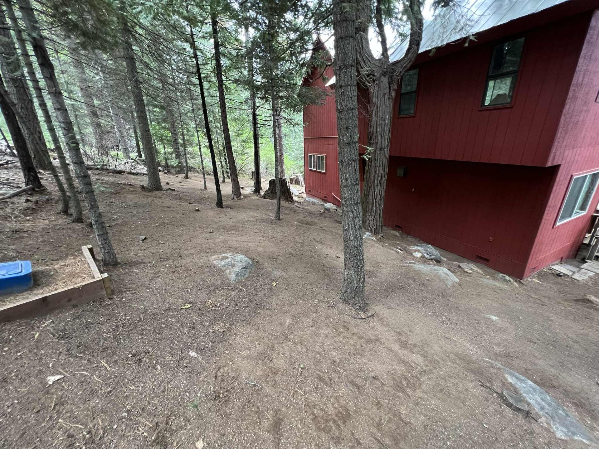 Red house on a wooded hillside. Brown ground with scattered trees and rocks. Blue storage box.