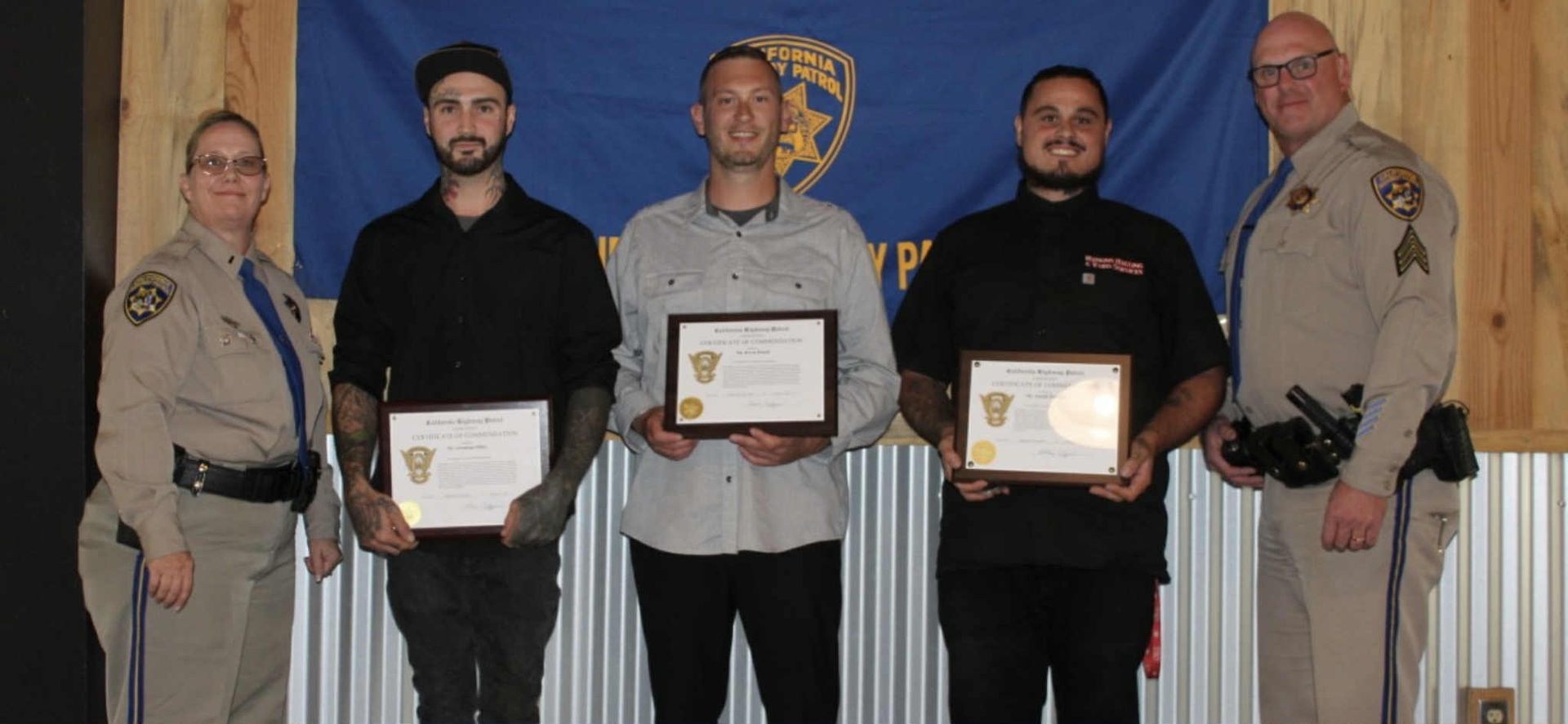 Five people holding certificates; two are in uniform in front of a blue banner.