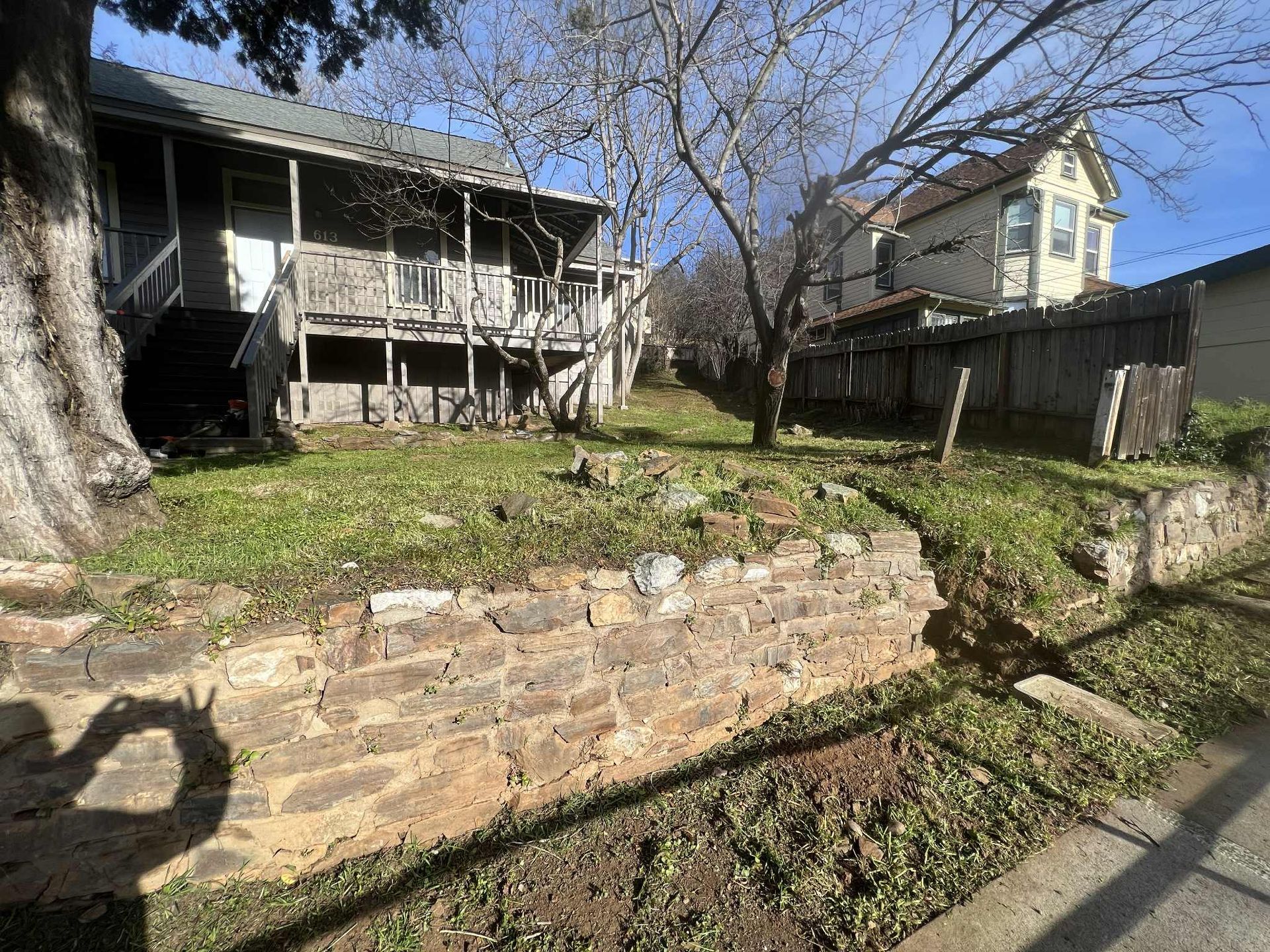 Stone retaining wall and houses with overgrown grass and trees.