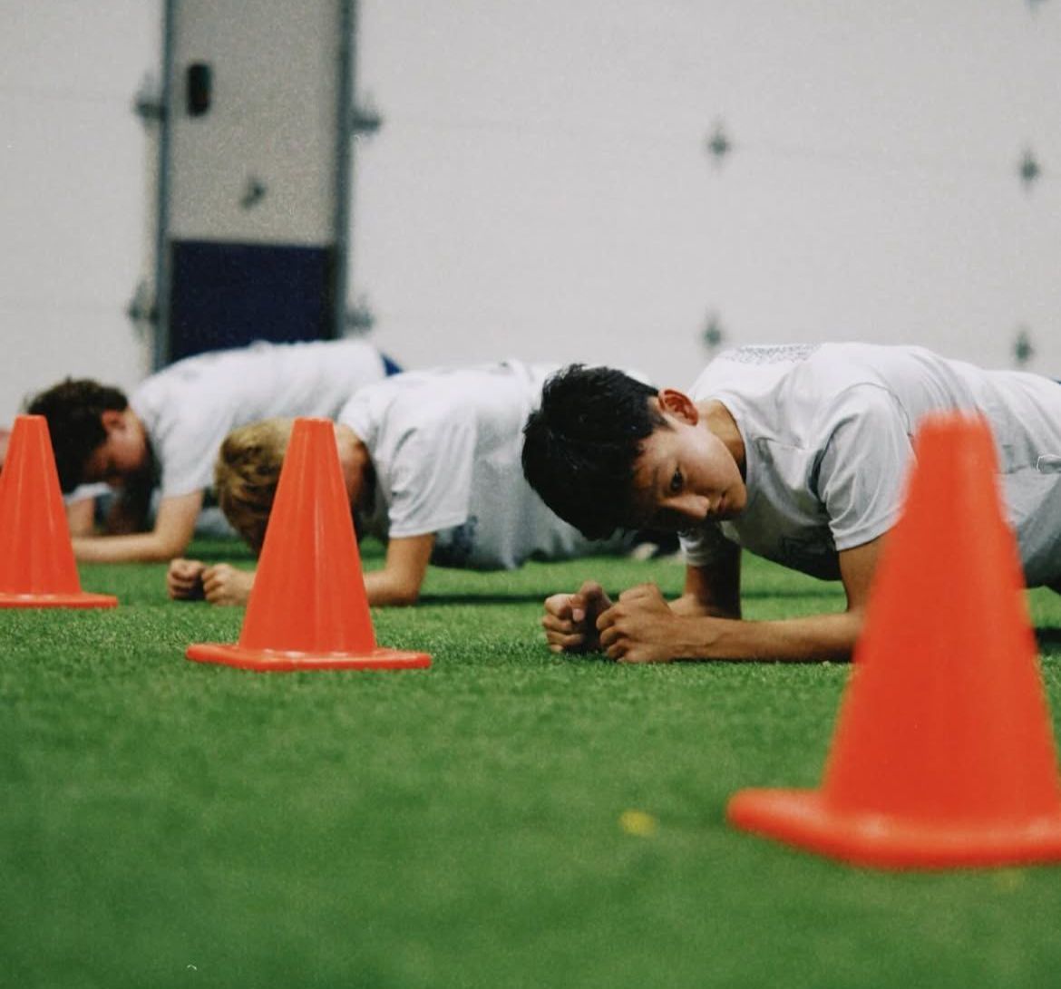 People doing plank exercises on green turf between orange cones indoors