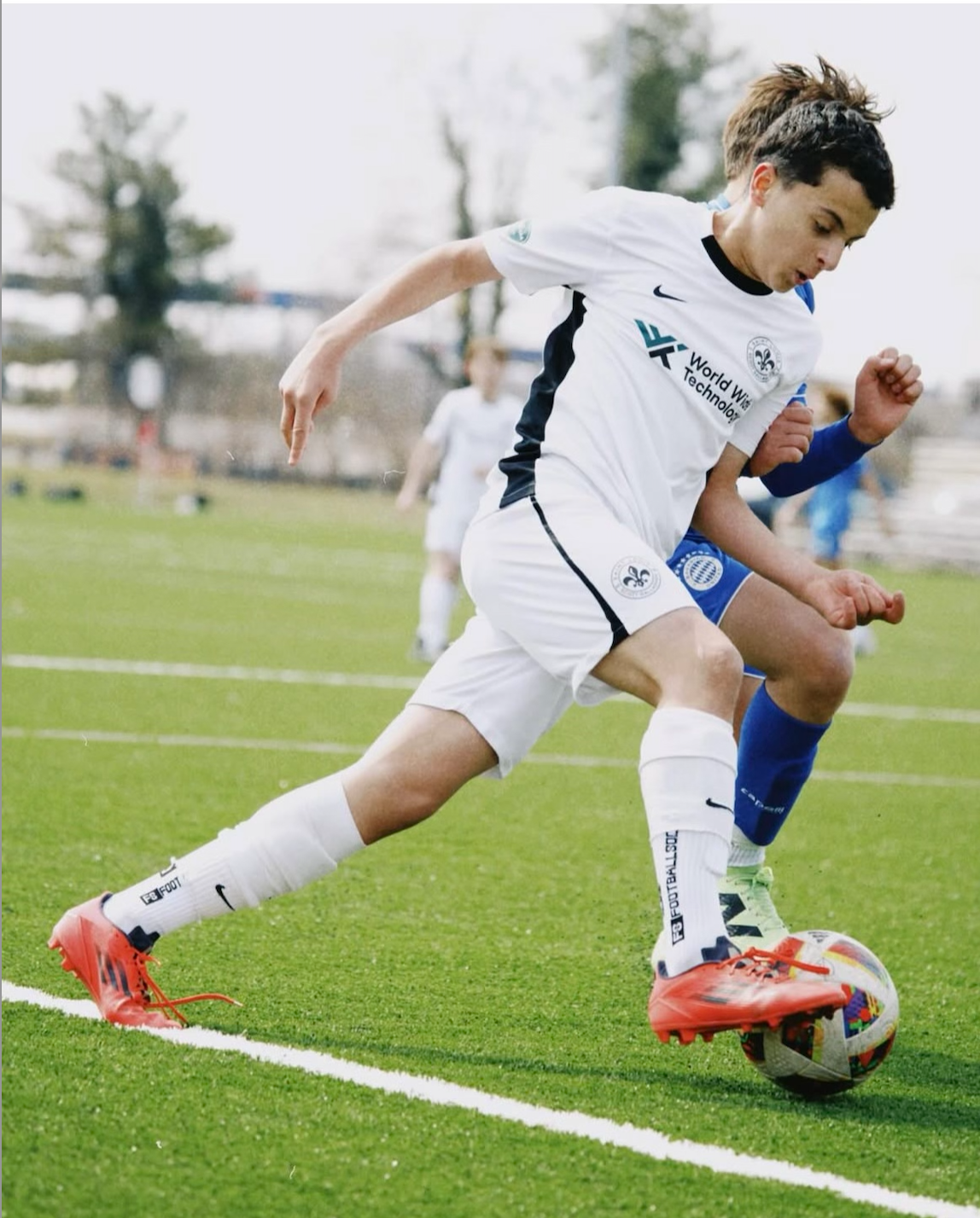 Two soccer players contesting the ball on a green field, one in white and one in blue.