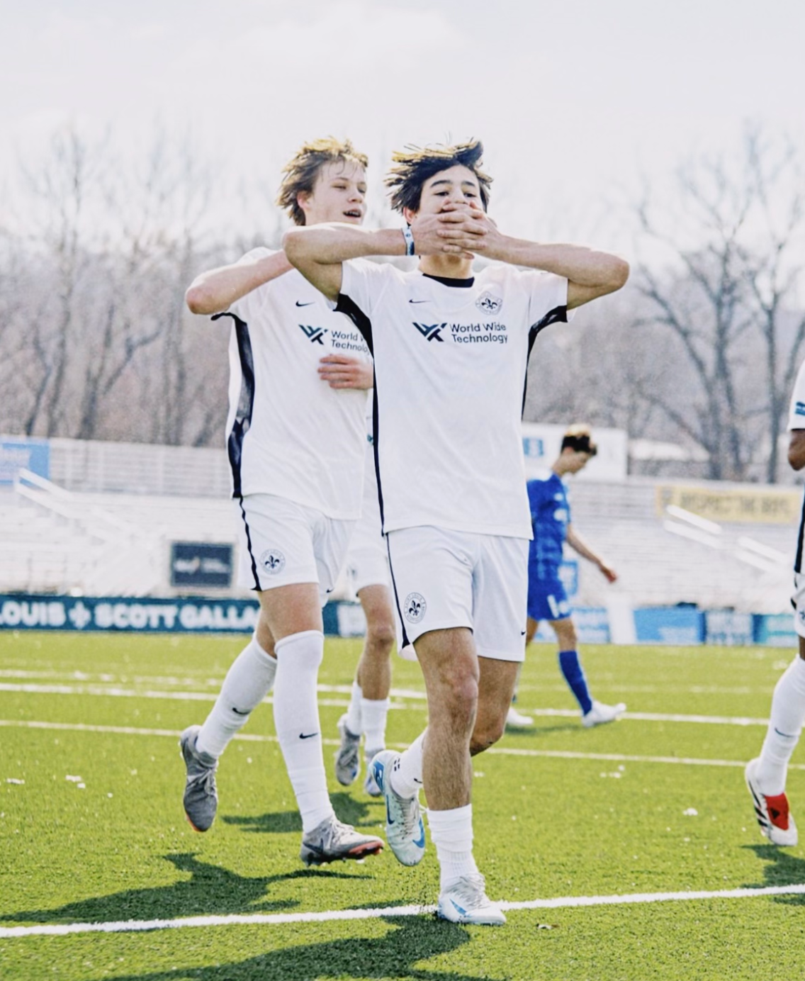 Two soccer players in white jerseys celebrate on the field with arms raised.