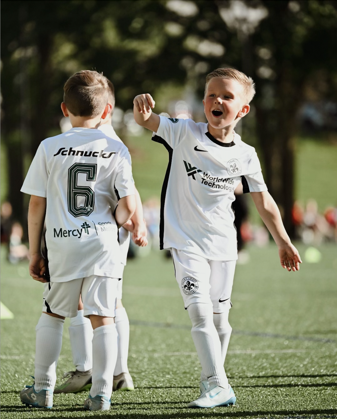 Two young soccer players in white uniforms celebrating on a grassy field.