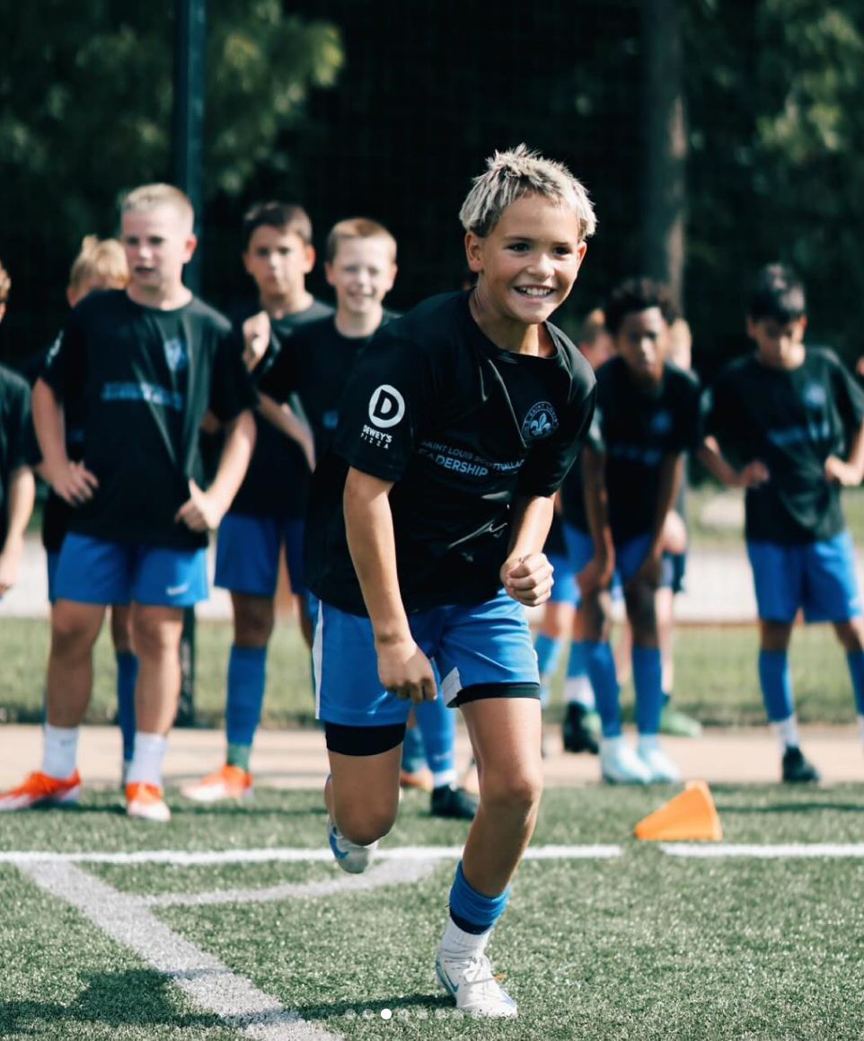 Children doing sprint drills on a turf field, one smiling runner in a black shirt and blue shorts in front.