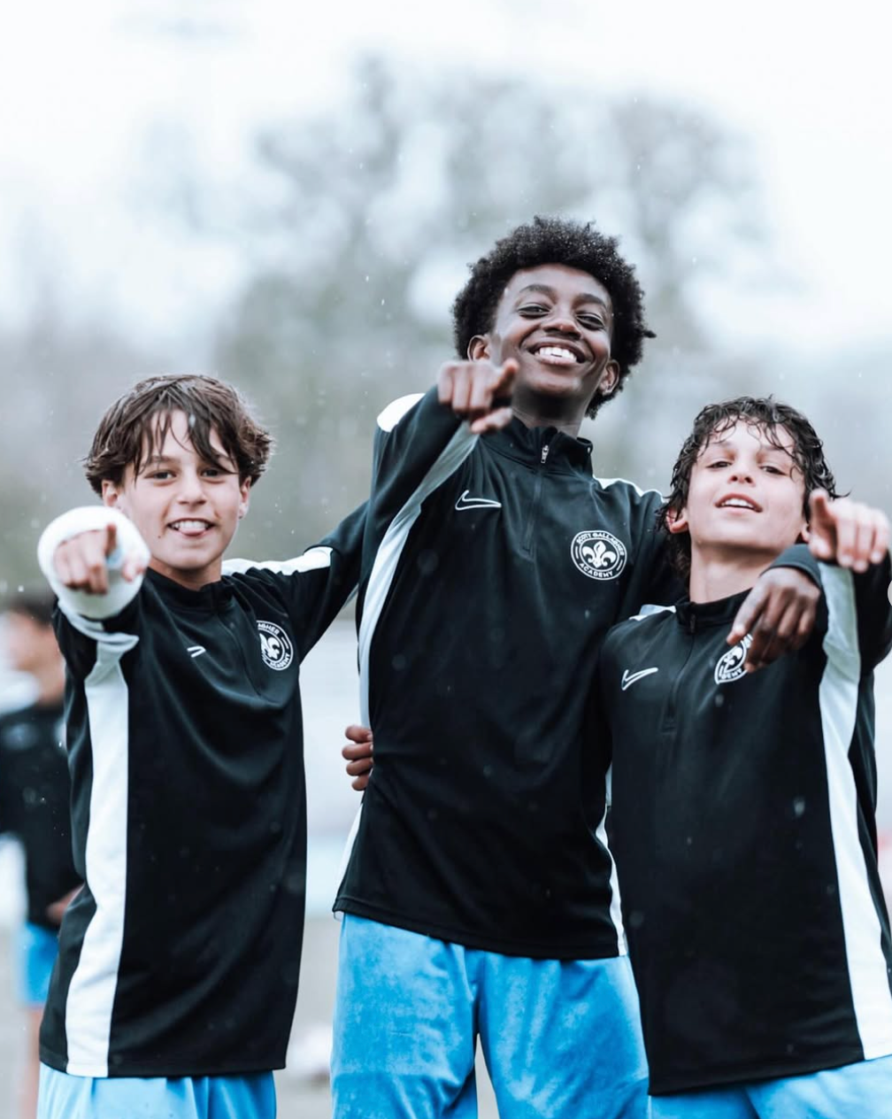 Three soccer teammates in black jerseys smile with arms around each other on a field.