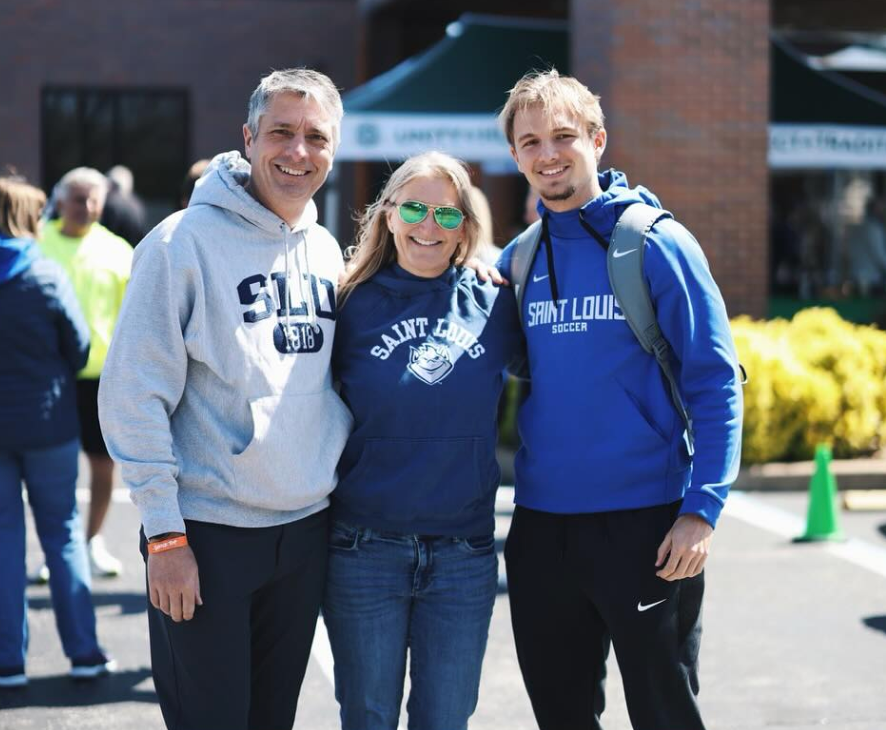Three people smiling outdoors in blue and gray Saint Louis hoodies on a sunny day