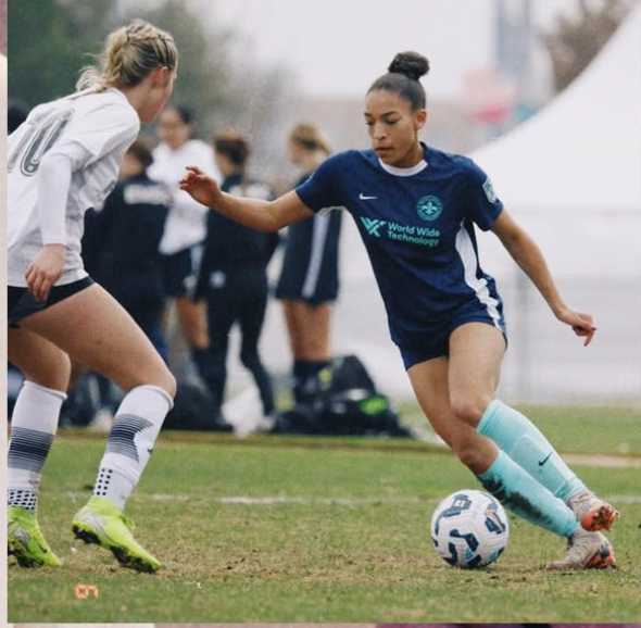 Two soccer players vie for the ball on a grass field, one in navy, one in white.