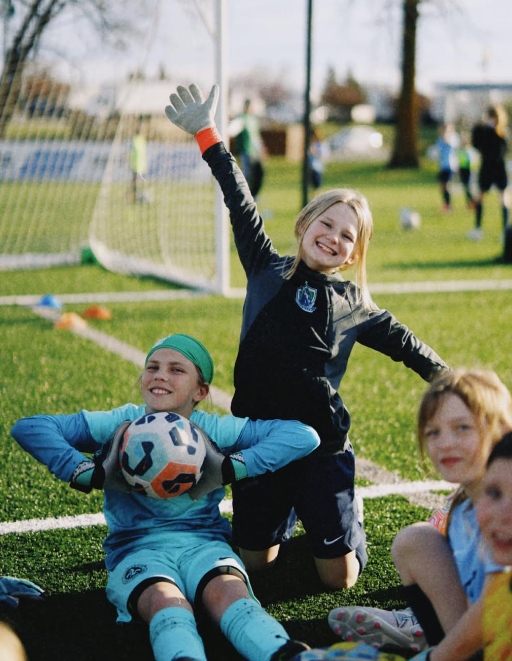 Children in soccer gear sit and cheer near a goal on a grassy field