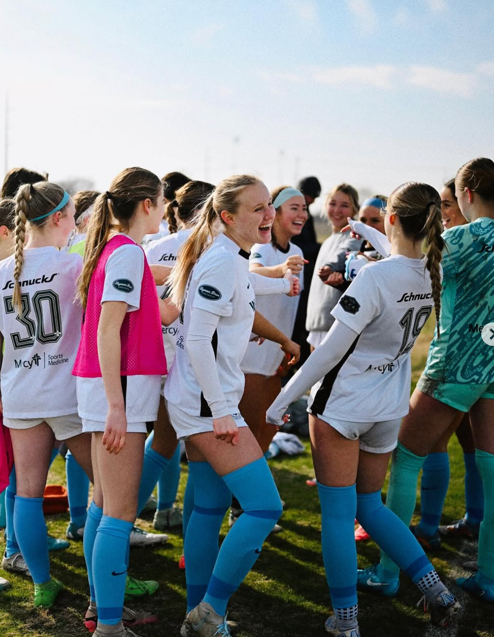 Soccer players in white and teal uniforms huddle on a field, smiling and chatting before play.