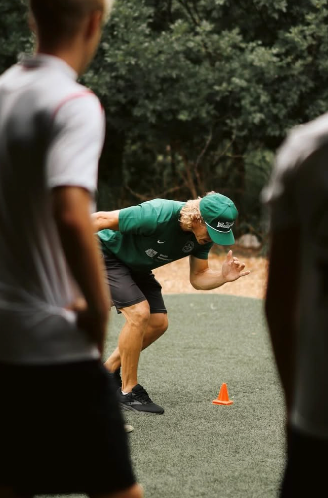 Man in green cap and shirt crouches for a drill on a grassy field, with an orange cone nearby and others blurred in foreground