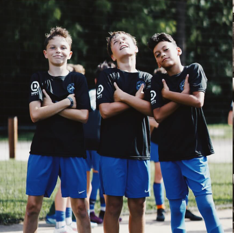 Three children in black jerseys and blue shorts stand with arms crossed on a soccer field, smiling.