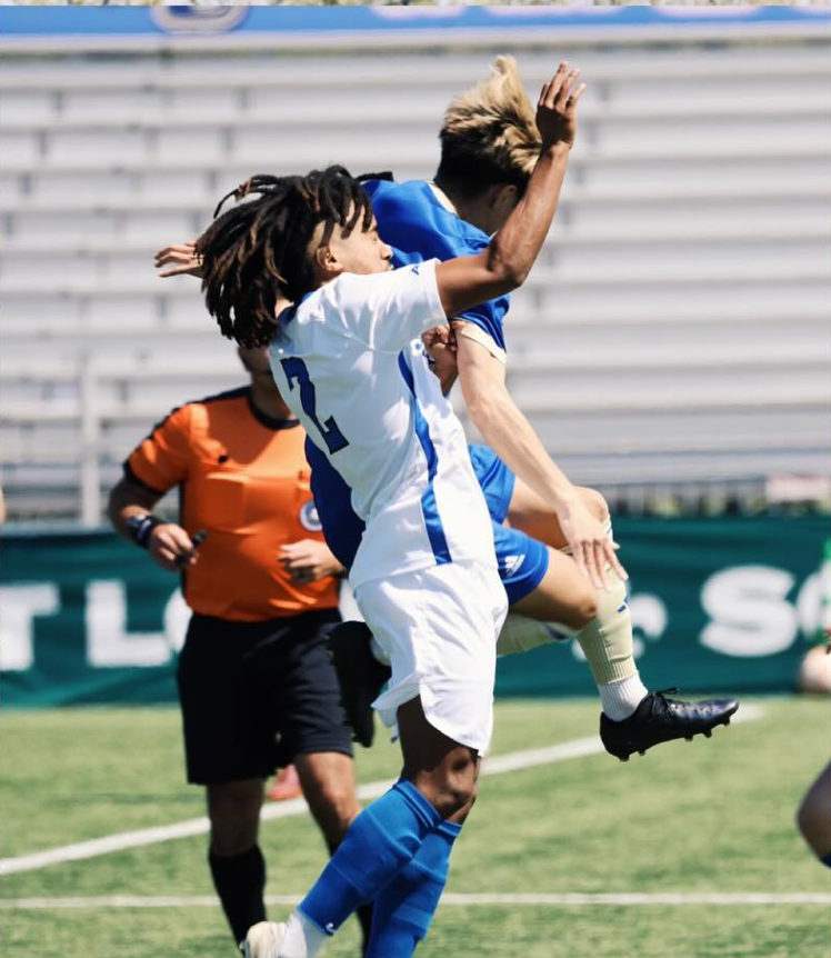 Two soccer players leap for a header during a match on a sunny field.