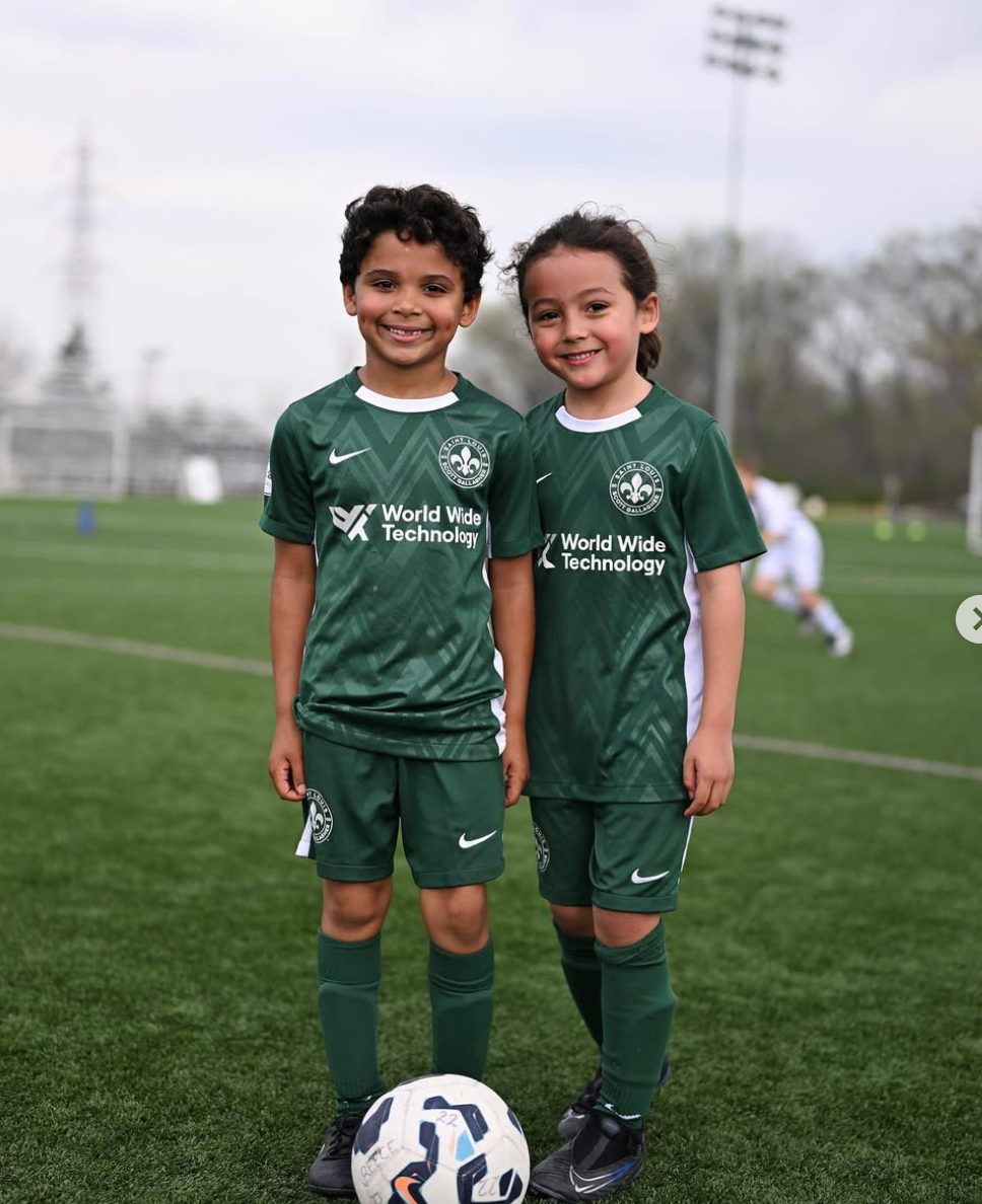 Two children in green soccer uniforms stand on a field with a soccer ball, smiling at the camera.