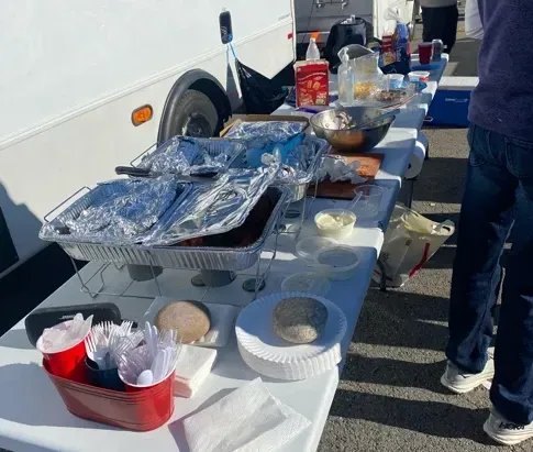 Outdoor buffet table beside a white truck, covered with foil trays, bowls, and drinks.