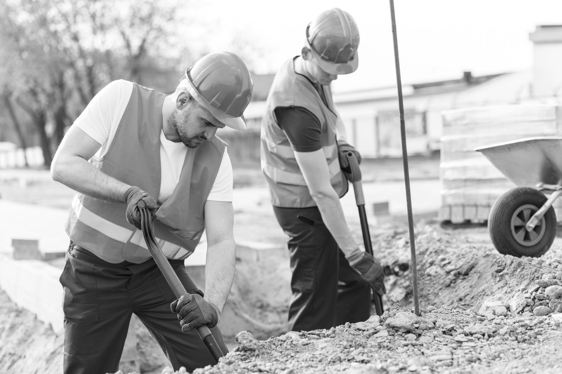 Two construction workers in orange vests and hard hats shoveling gravel at a roadwork site