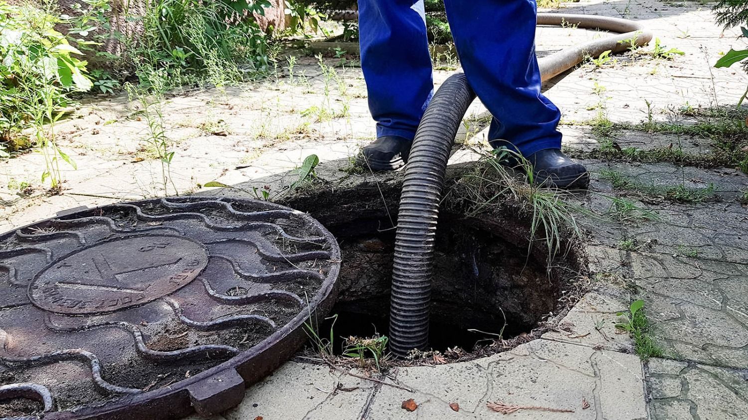 Worker in blue pants using a hose to clear a sewer manhole on a sidewalk
