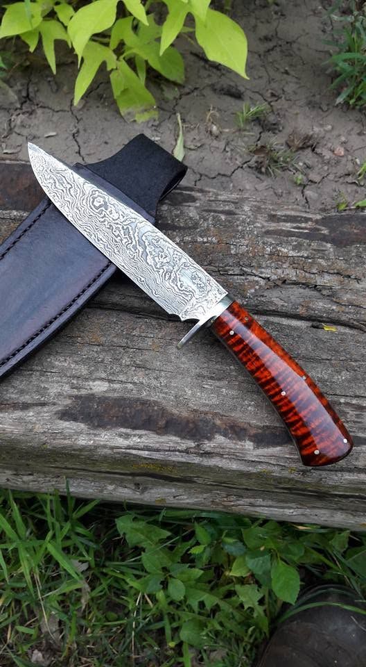 Decorative knife with red striped handle resting on a wooden log beside a black sheath outdoors