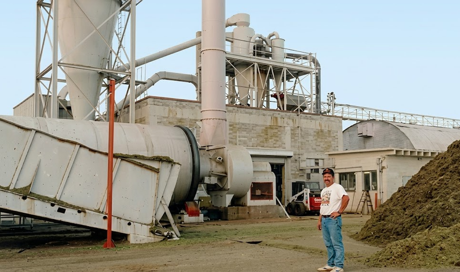 Industrial facility with a large metal silo and machinery, a man standing beside a mound of material.
