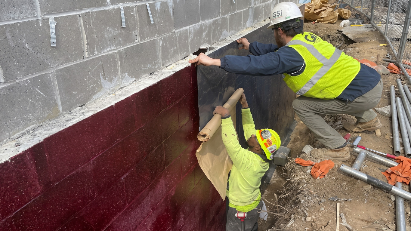 Two workers installing a dark red wall membrane on a cinder block wall at a construction site