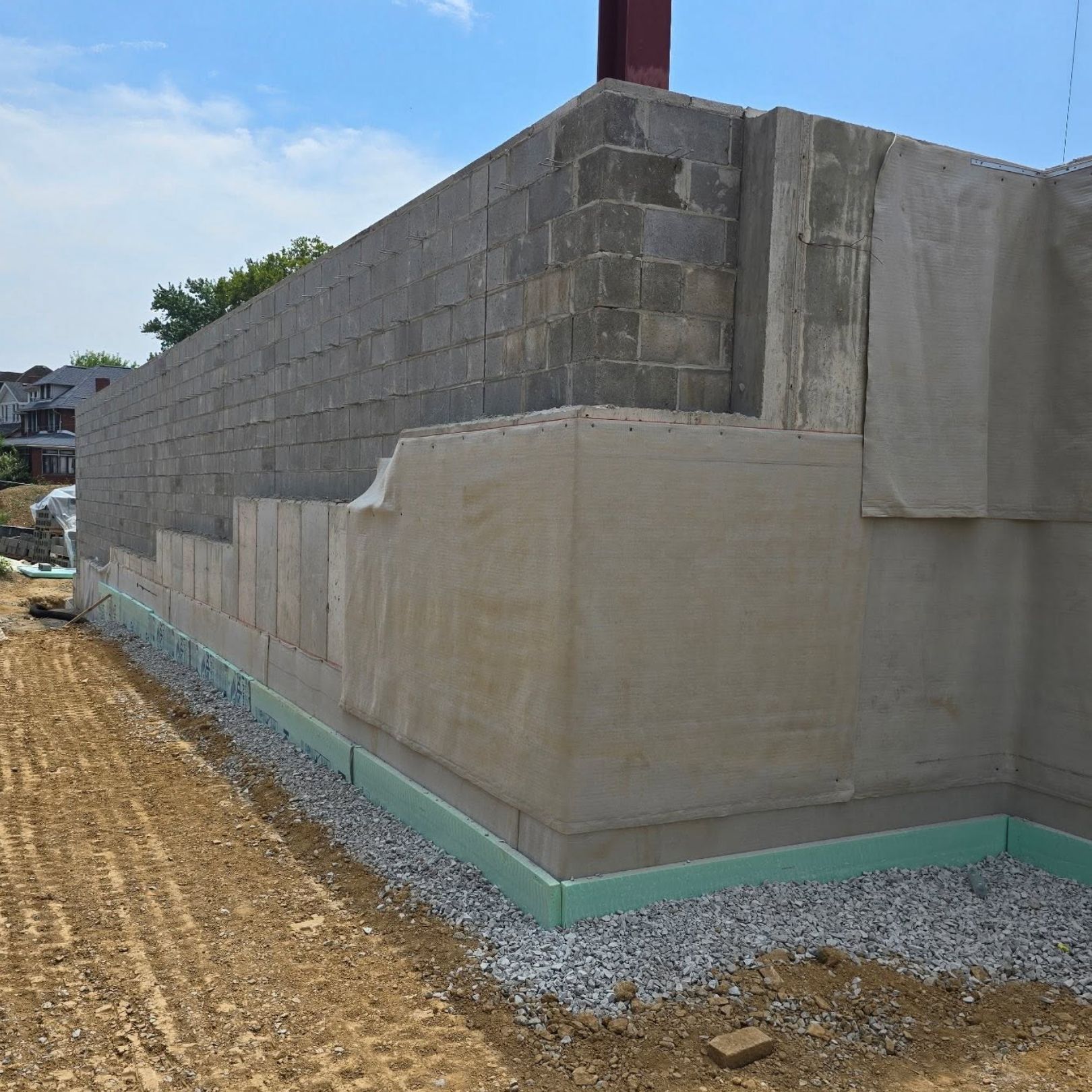 Concrete foundation wall under construction beside a dirt lot, with gray cinder blocks and green base waterproofing