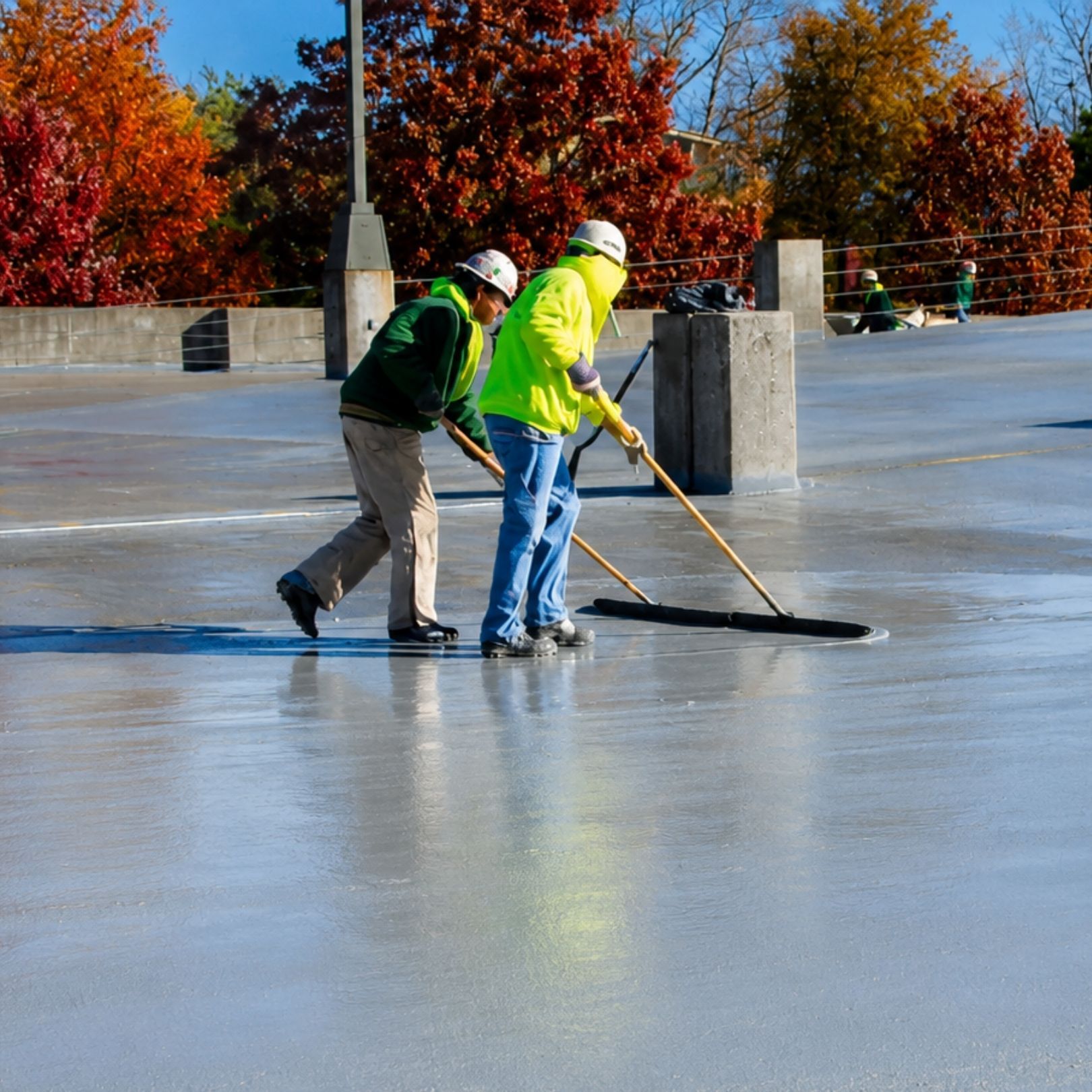 Two workers in hard hats smoothing wet concrete on a rooftop under autumn trees