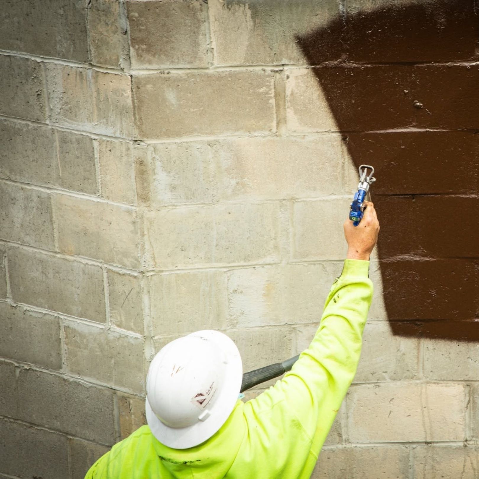 Worker in white hard hat painting a concrete wall corner brown with a brush.
