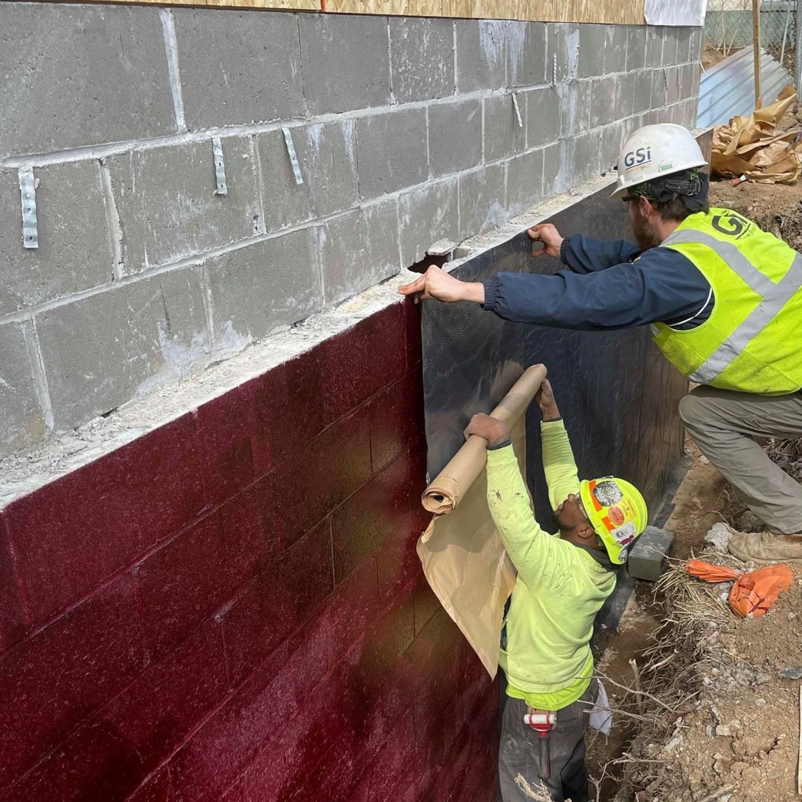 Construction workers installing maroon wall panels on a gray cinderblock foundation at a worksite