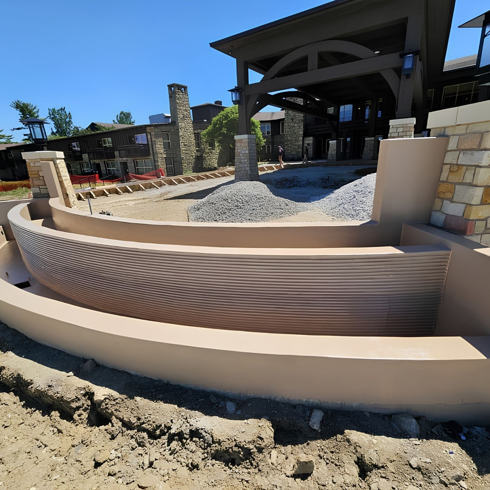 Curved tan retaining wall beside gravel landscaping and a covered outdoor pavilion in bright sunlight