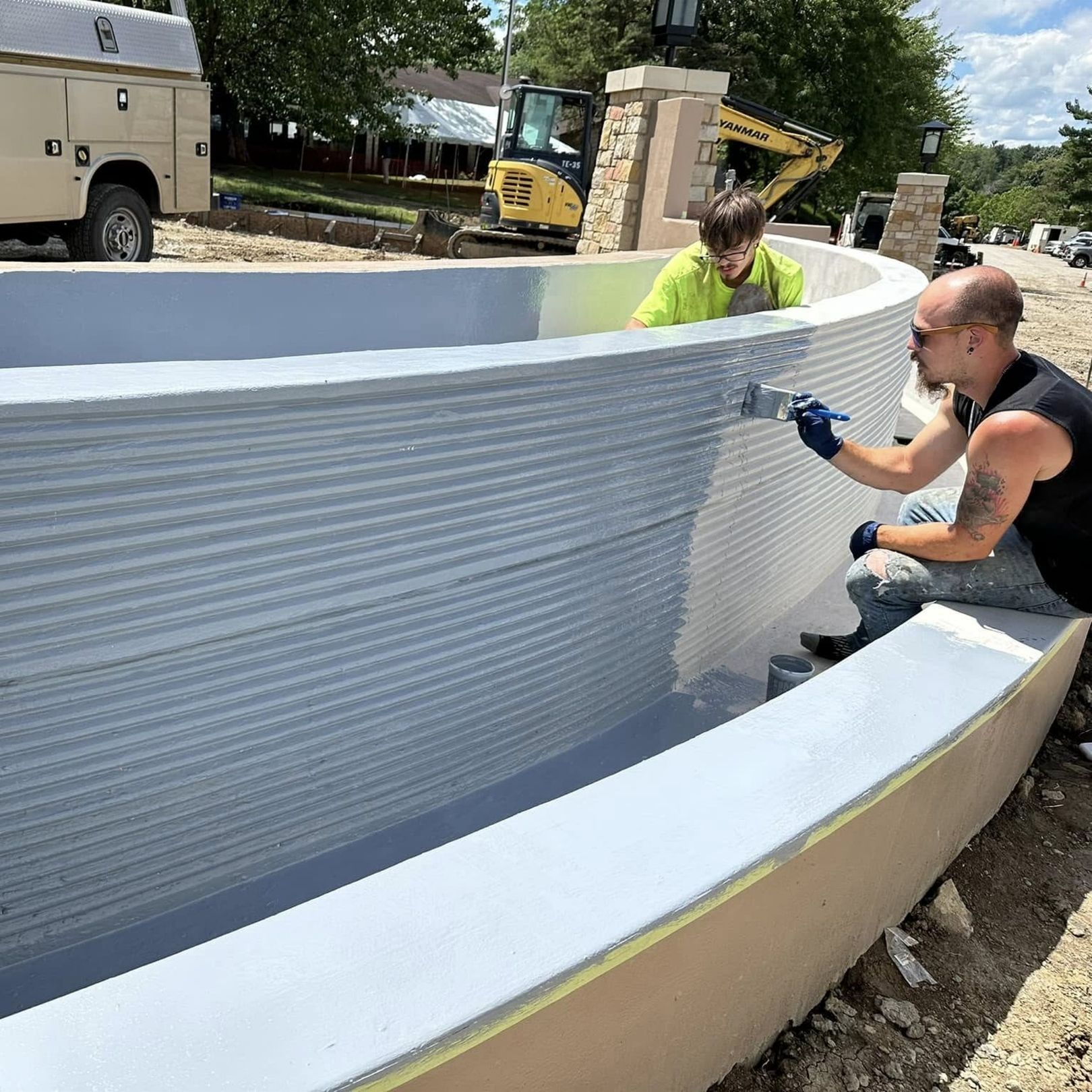 Two workers painting a curved outdoor concrete wall white at a construction site