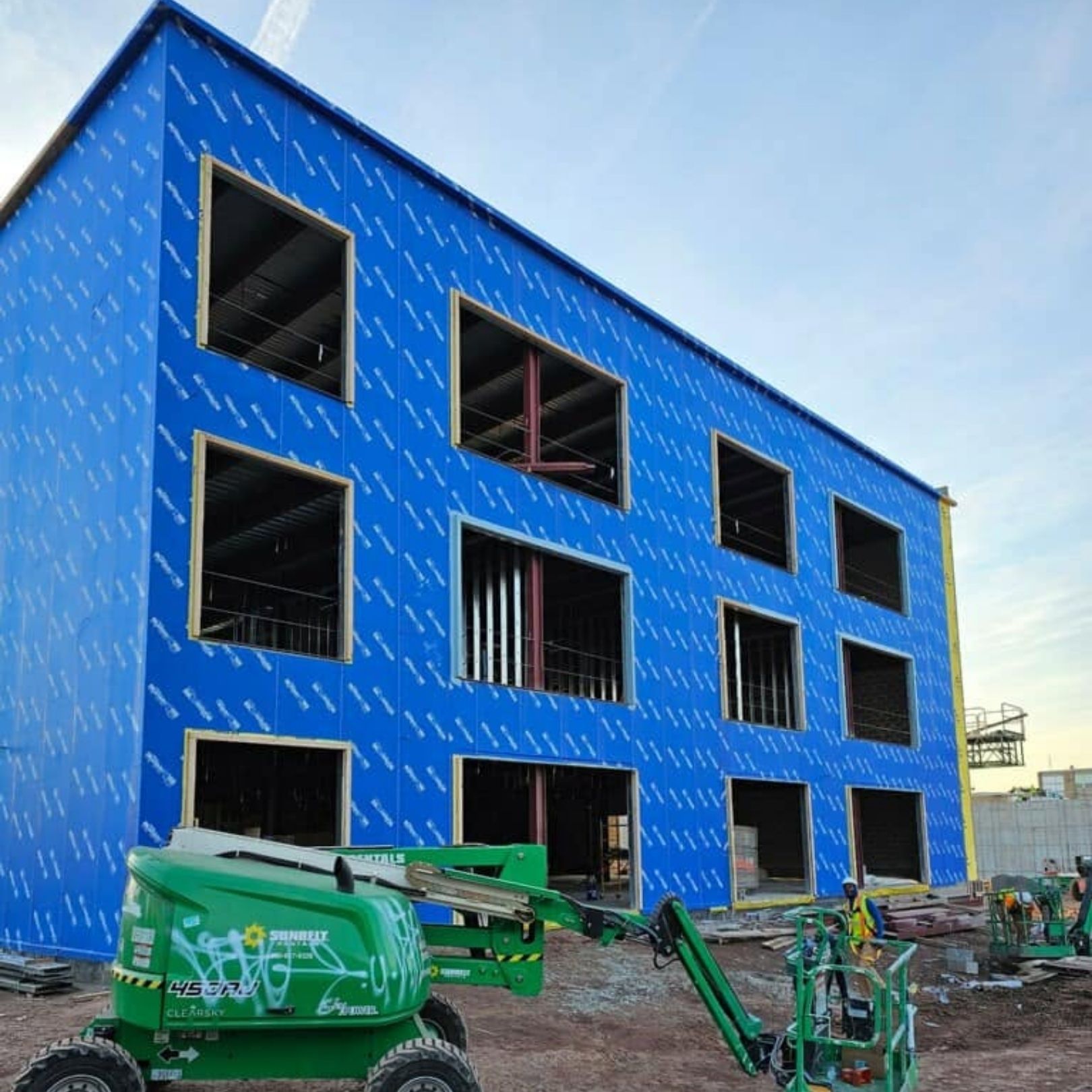 Blue building under construction with large window openings and green construction lifts in front
