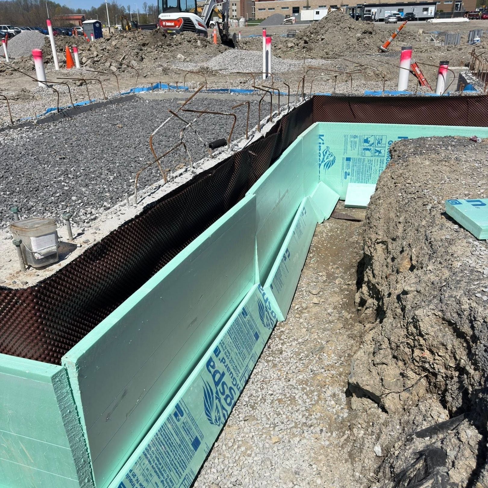 Construction site with green foam foundation walls, gravel trench, and exposed rebar under bright daylight