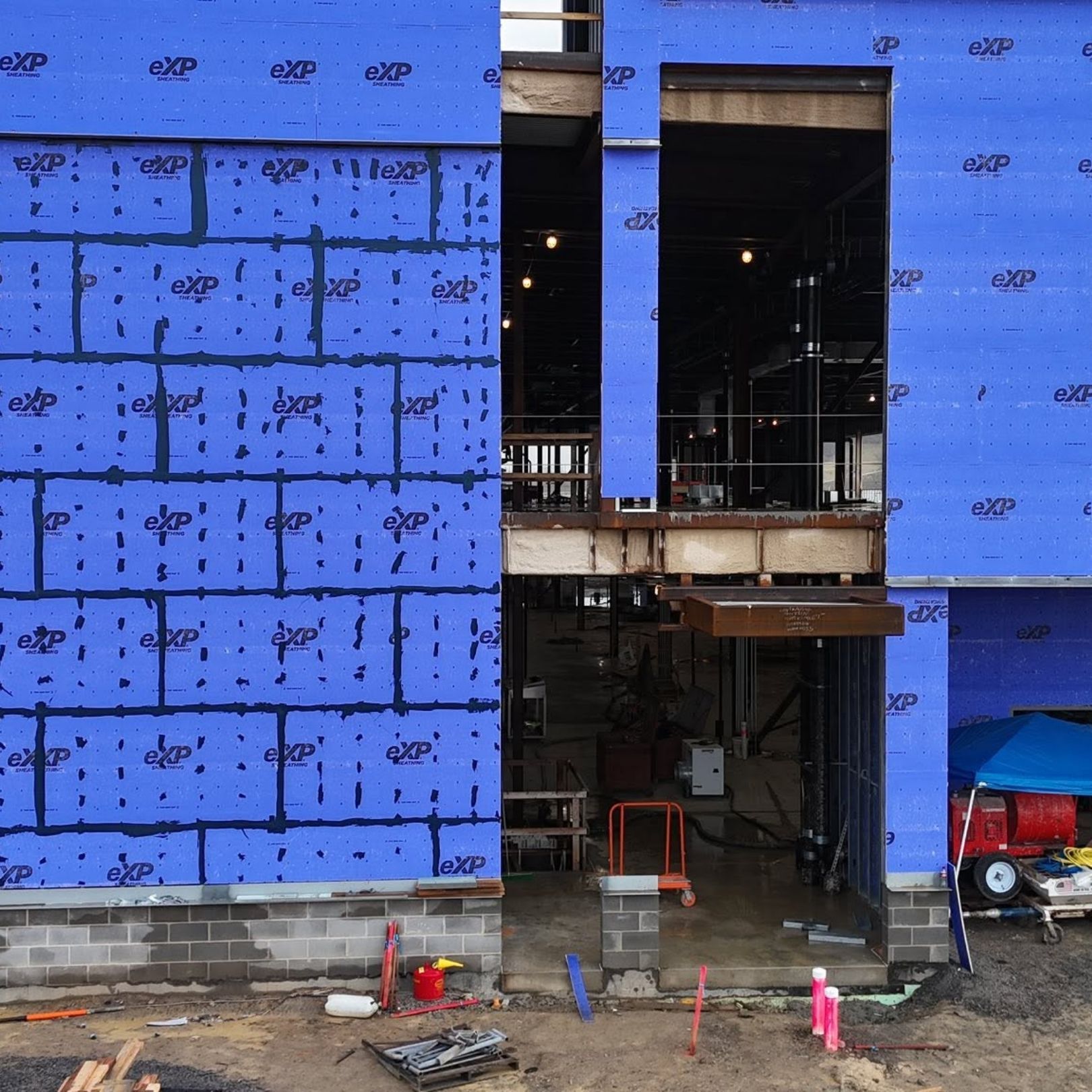 Blue-sided building under construction with open doorway, exposed framing, and construction equipment at ground level
