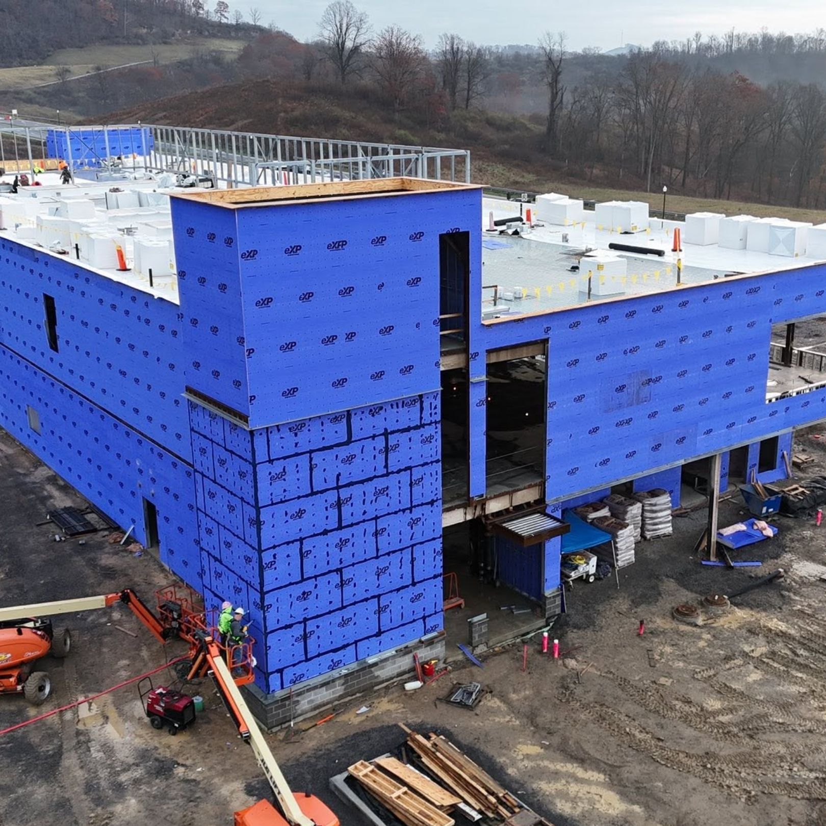 Blue-wrapped building under construction with scaffolding and equipment on a muddy site