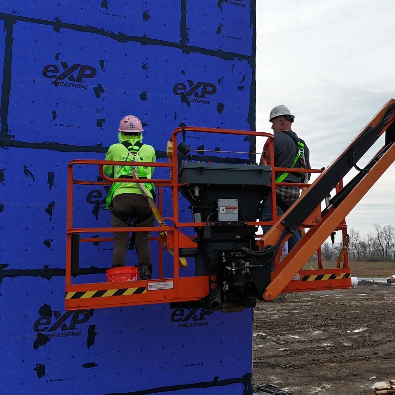 Two workers on an orange lift install blue wall panels at a construction site.