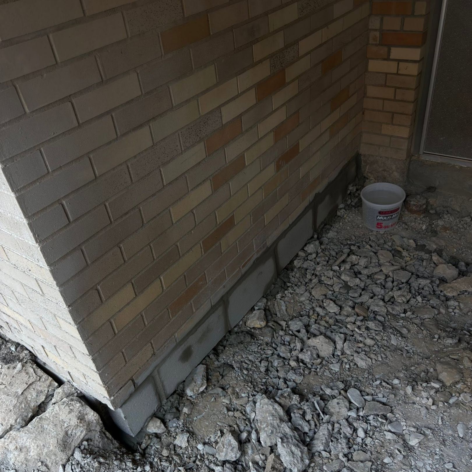 Corner of a brick house with damaged foundation and gravel ground, beside a white bucket near a door.