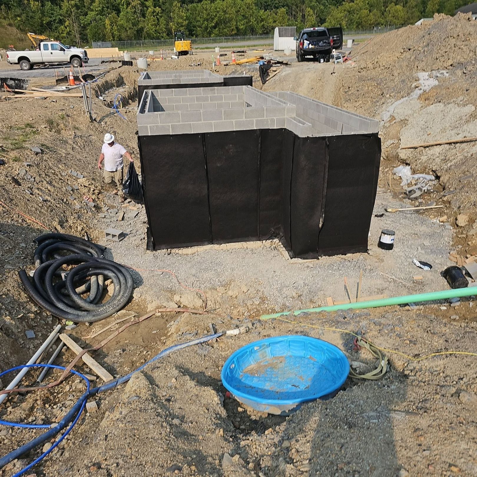 Construction site with a black foundation structure, pipes, dirt piles, and workers in the background