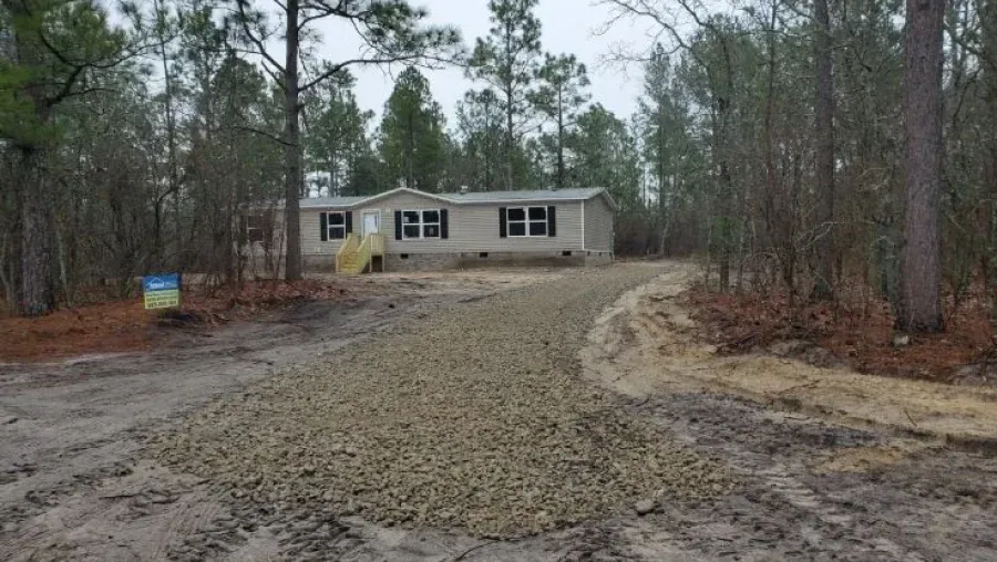 A beige manufactured home sits in a wooded lot with a newly laid gravel driveway leading to the front stairs.