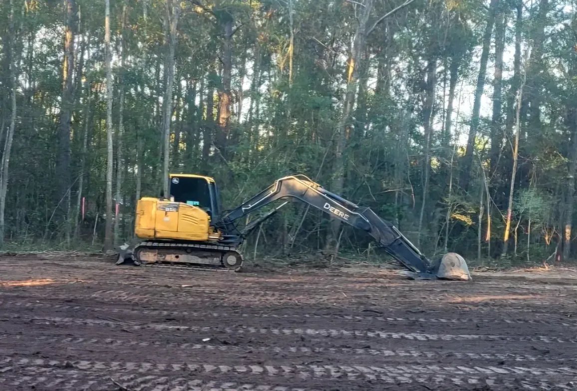 A yellow and black excavator sits on a cleared, muddy lot in front of a dense forest.