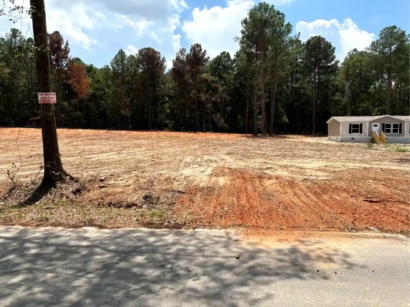 A yellow and black excavator sits on a cleared, muddy lot in front of a dense forest.
