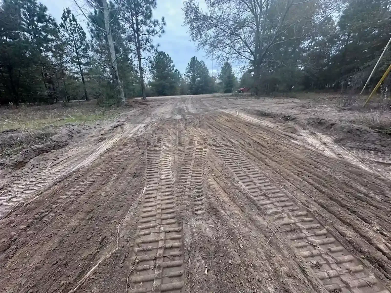 A wide, unpaved dirt path through a wooded area featuring heavy tire tracks from construction equipment.