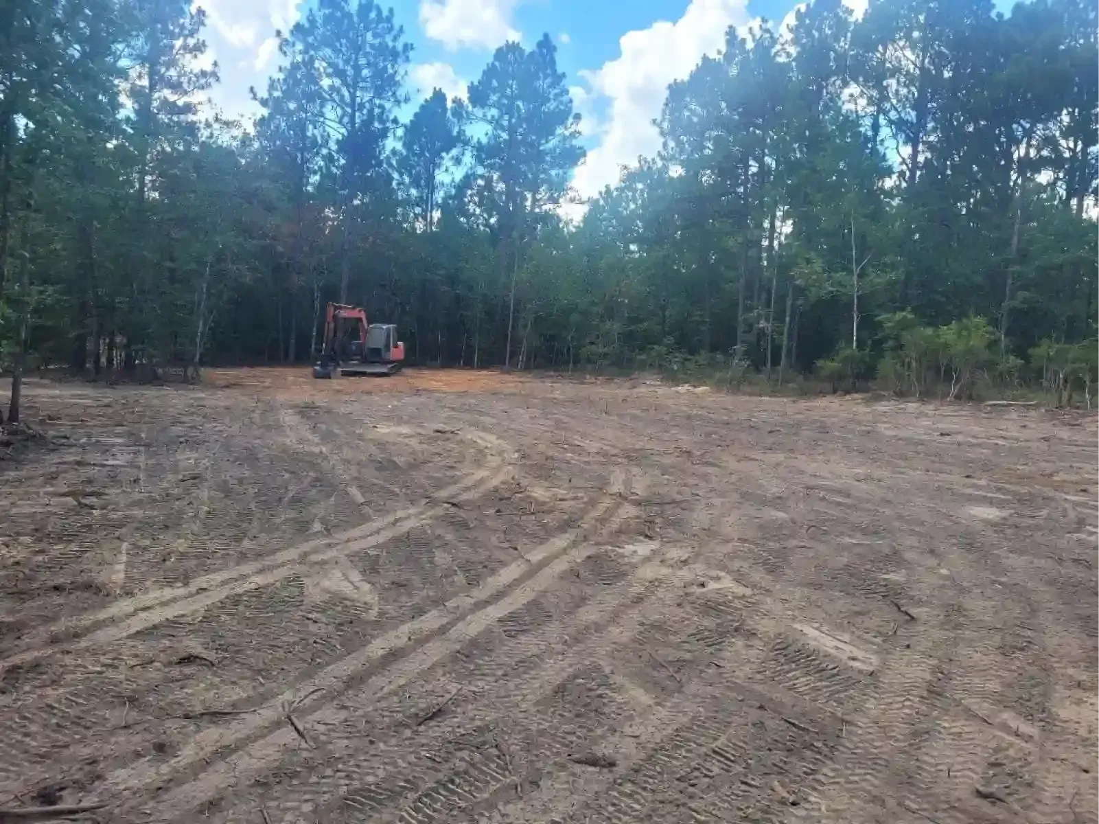 A wide, cleared construction site with dark soil, tire tracks in the foreground, and a small building under a blue sky.